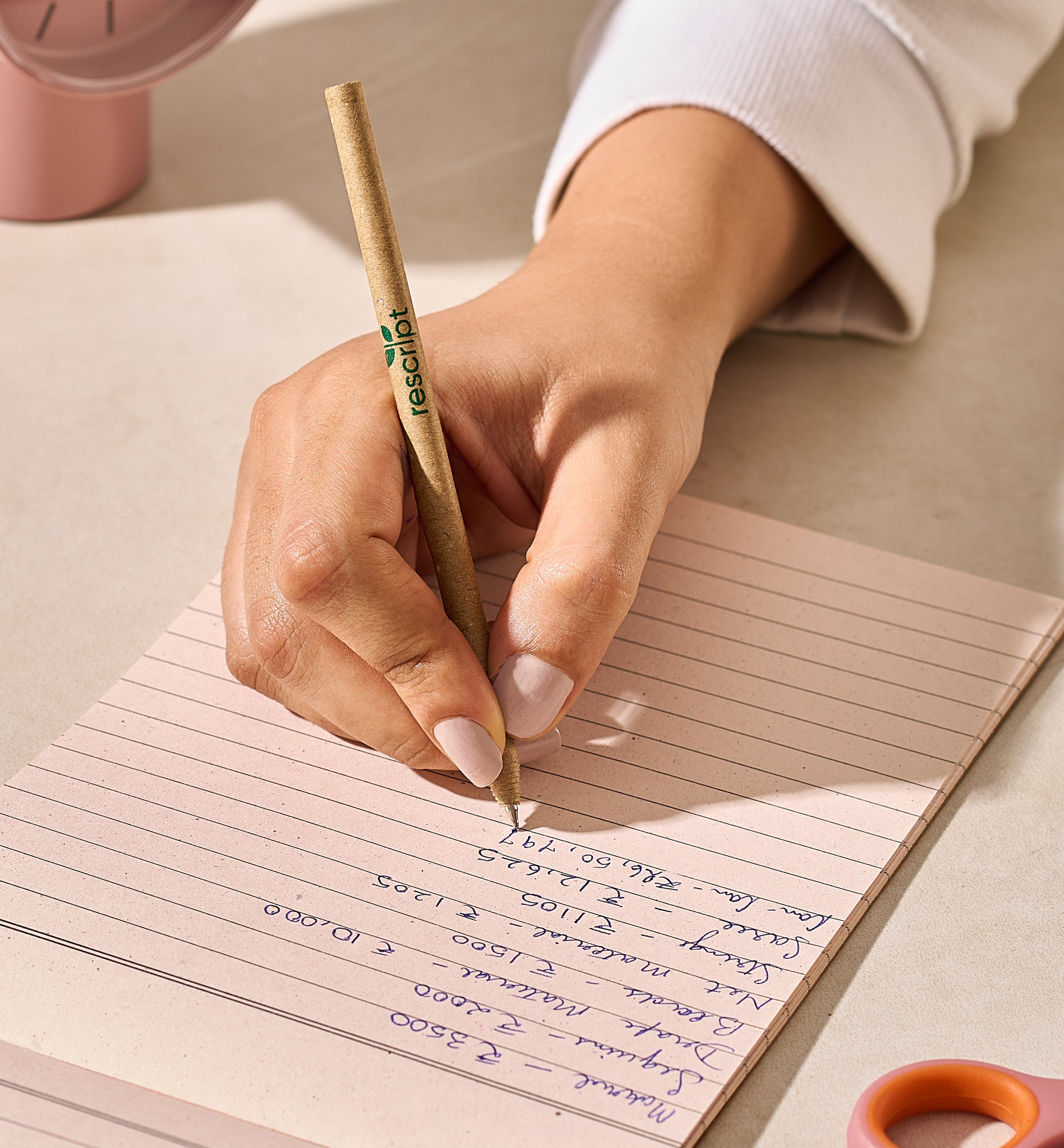 Hand writing on lined paper with a pen, pink clock and scissors in the background