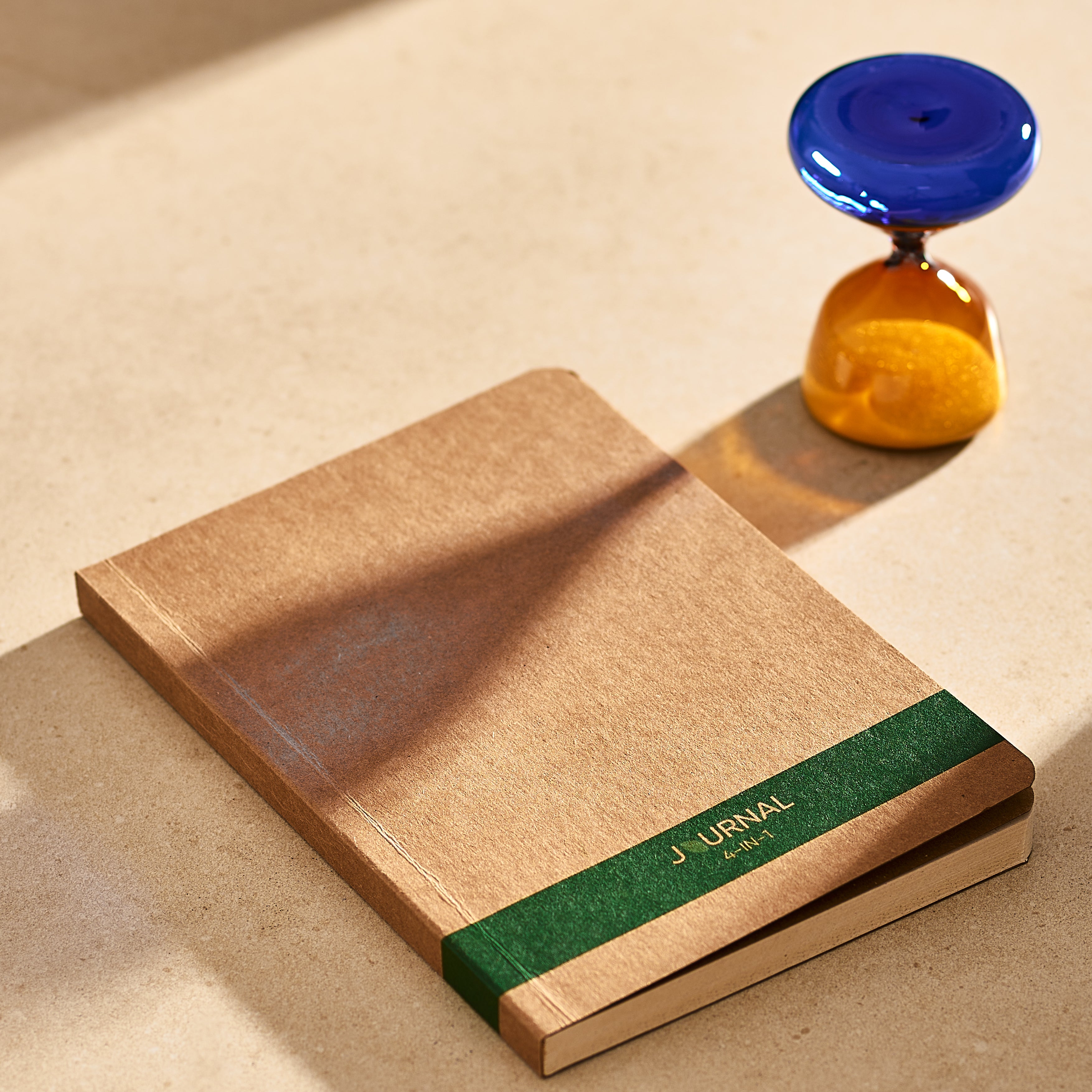 Eco-friendly A5 journal with recycled paper and a green spine, placed next to a blue and amber hourglass.